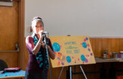 A young indigenous woman with her hair in a bun speaks in a microphone at a well-lit room. A sign behind her says "What Does Wellness Feel Like?"