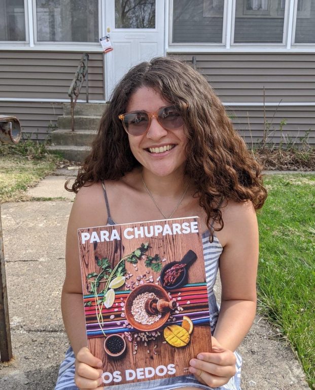 A Latinx woman with curly hair and sunglasses holds a copy of the cookbook.
