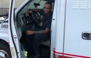 A young African American man sits in the driver's seat in an ambulance and smiles.
