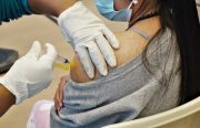 A closeup image of a young woman receiving a shot on her shoulder, with the person administering wearing latex gloves.