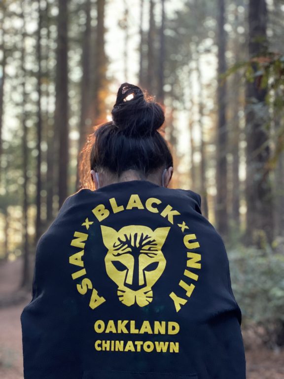 A woman in a forest wearing an Asian Black Unity black hoodie with her back facing camera