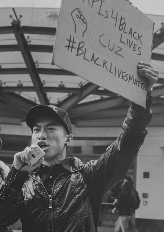 An image of an Asian American man speaking into a mic and holding a sign that says Asians4BlackLives cuz Black Lives Matter