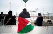 A Palestinian flag with red, green, and white triangles is wrapped around a person's back, with the Port of Oakland's cranes in the background