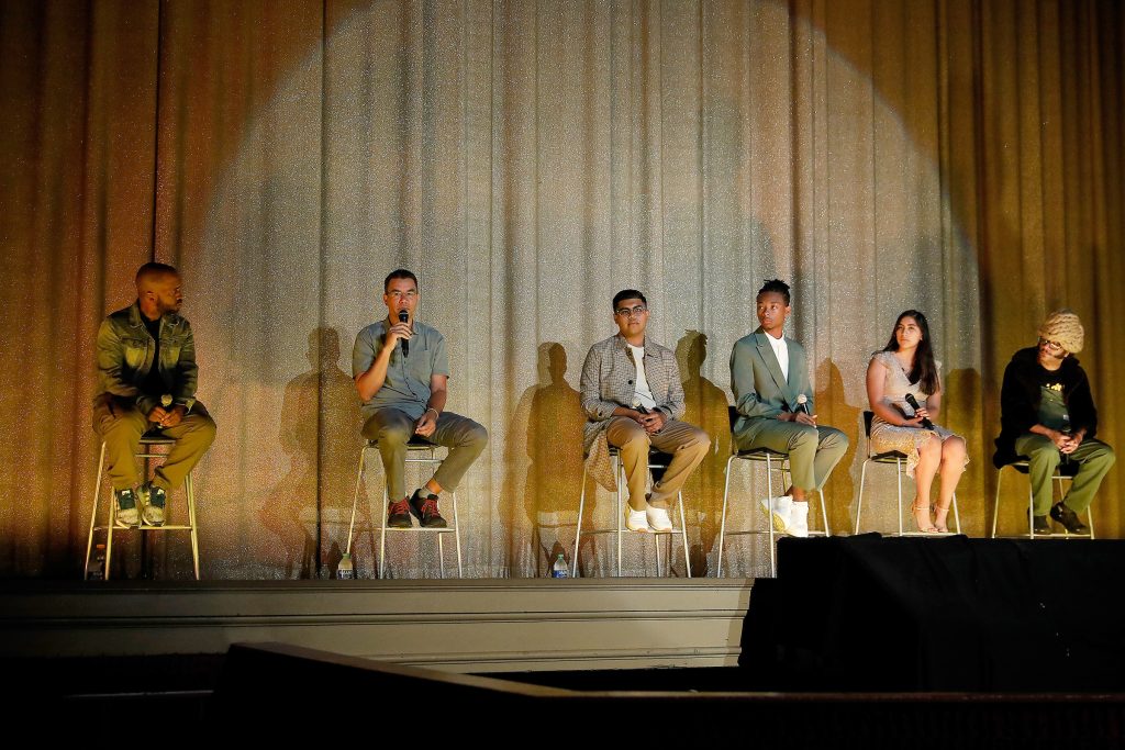 A stage lit up in a dark theatre with a diverse group of youth and adults sitting on stage in front of a gold colored curtain at the Grand Lake Theatre in Oakland.