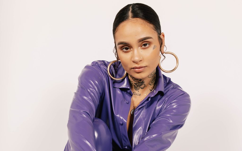 A biracial woman with slicked back hair and tattoos on her face is wearing a purple vinyl jacket