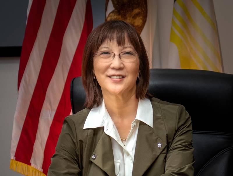 An Asian American woman with bangs and glasses poses for photo in front of American flag