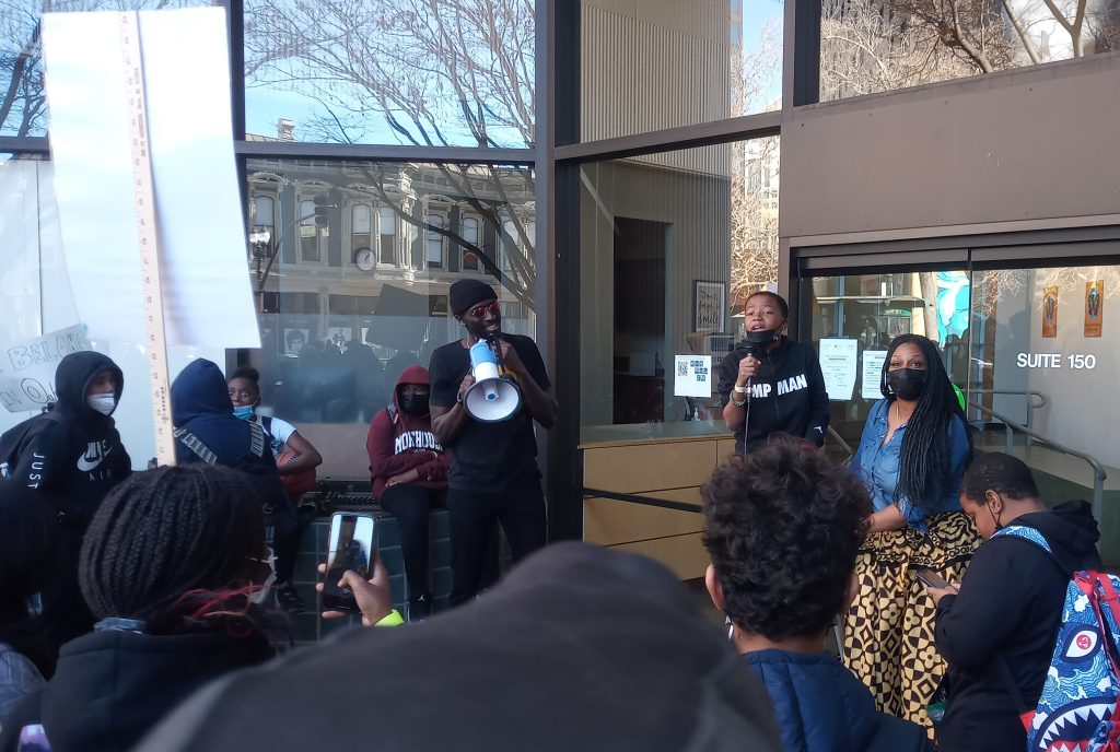 A young Black child speaks to a group, and on his left, a Black man holding a megaphone looks at him in support.