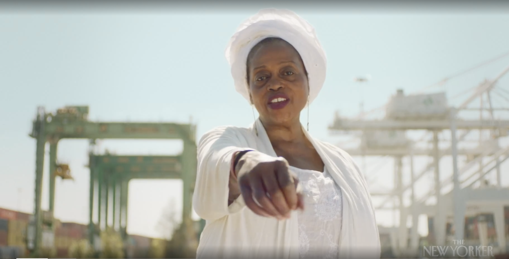 An African American woman wearing all white and a white hair wrap rolls dice towards camera. In the background is Oakland's cranes and shipyards.