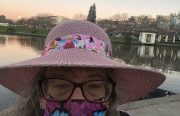 A woman wearing a purple mask, pink hat, and glasses takes a selfie in front of Lake Merritt.