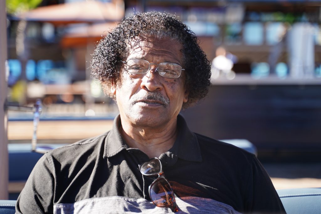 An African American man with curly hair and glasses sits outside on a sunny day