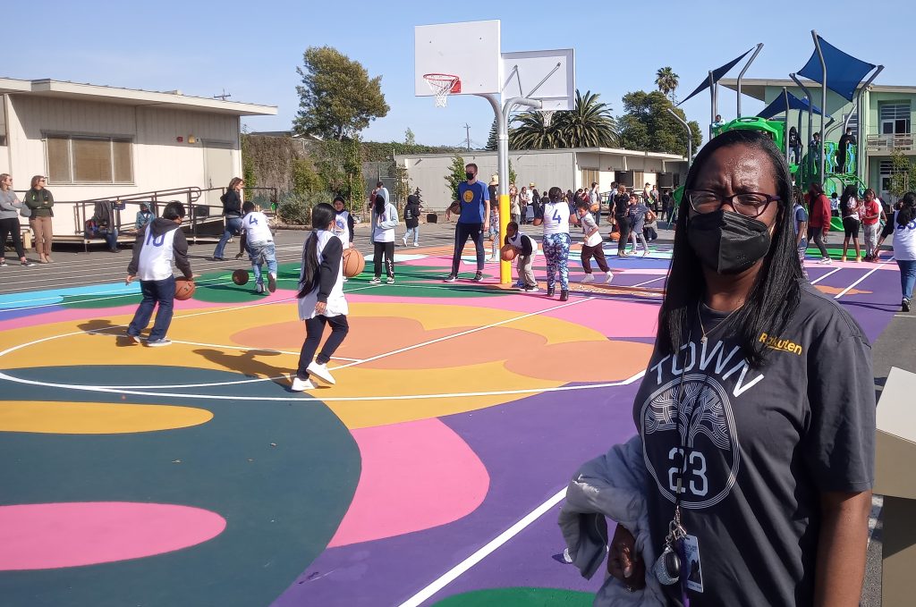 Am African American woman teacher stands near colorful outdoor basketball court with kids playing ball in the background