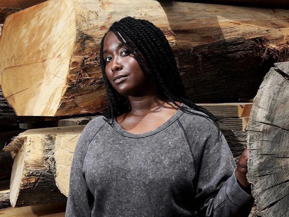 A black women with braids wearing a grey sweater poses in front of large pieces of logs and wood