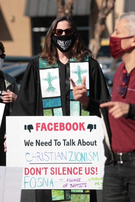 A woman wearing a black face mask and sunglasses holds sign that says "Facebook: we need to talk about Christian Zionism"
