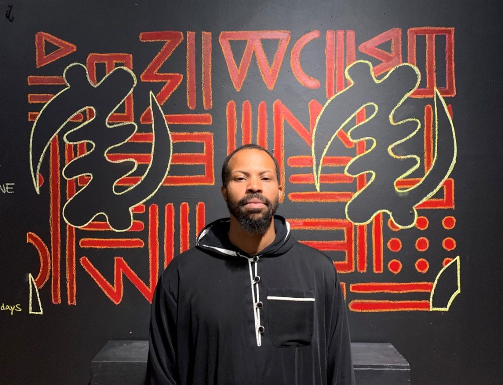 An African American man with short hair in front of red and black mural with Chinese words